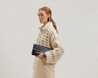 Woman wearing a beige outfit with a textured sweater and long skirt, holding a black clutch against a plain background.