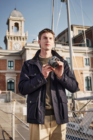 male model with blond hair, a blue modern down jacket with large front pockets, a dark green jumper and beige large trousers, with photocamera on hand