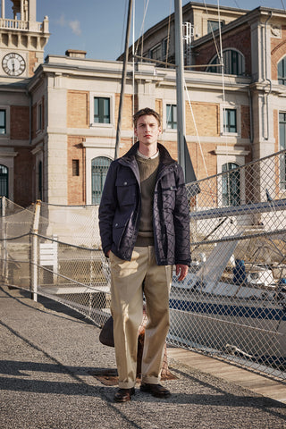 male model with blond hair, a blue modern down jacket with large front pockets, a dark green jumper and beige large trousers near the port