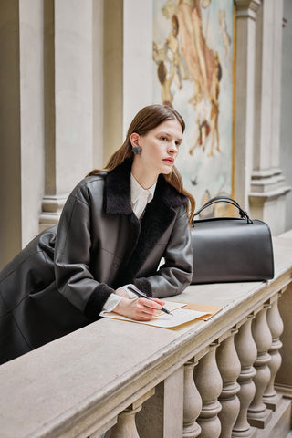 woman writing with a pen in an old palace wearing a dark brown sheepskin, with a white shirt, earrings and a brown trunk bag