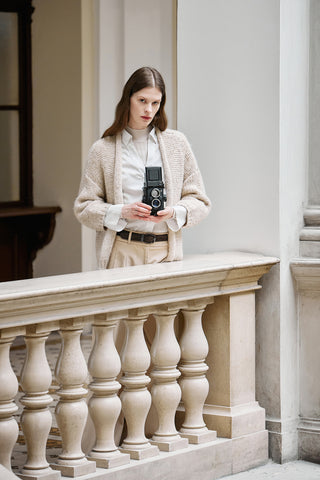woman wearing a beige tidy turtleneck sweater, a white shirt and a cardigan without bottoms, taking a picture with a vintage camera on the internal terrace of an old palace in trieste