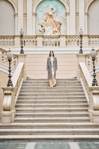 woman with a grey and brown long coat descending the stairs in an old palace with an afresco on the back