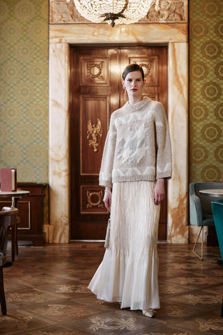 Woman in a white sweater and skirt standing in an elegant room with patterned wallpaper and wooden door.