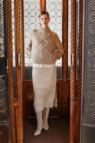 Woman wearing a beige knitted sweater and skirt standing in front of a decorative mirror.