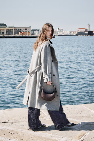 woman walking in front of the sea port wearing a grey long wool coat and blue fil coupé trousers and a brown moon bag