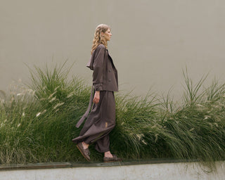 Woman in a long, dark outfit walking through tall grass with a neutral background
